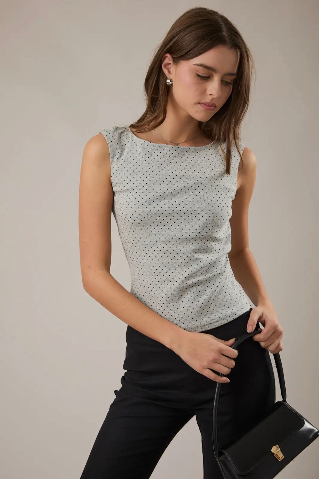 Woman wearing a sleeveless polka dot top and holding a black handbag against a neutral background